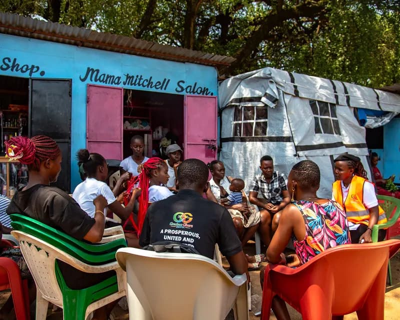 Young people, caregivers and Gracious Givers Kenya team members sit in a circle of plastic chairs for a community dialogue outside brightly painted market kiosks, one mother holding her baby.