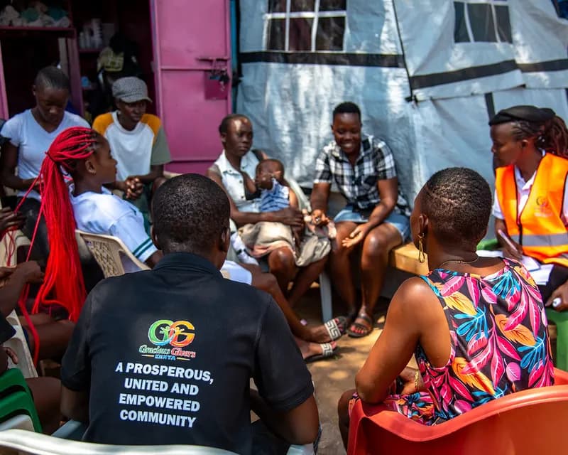 Caregivers, adolescents and young people sit in a close circle of plastic chairs outside a market kiosk in Nyando, one mother holding her baby, with a Gracious Givers Kenya facilitator in an orange vest listening at the edge of the group.