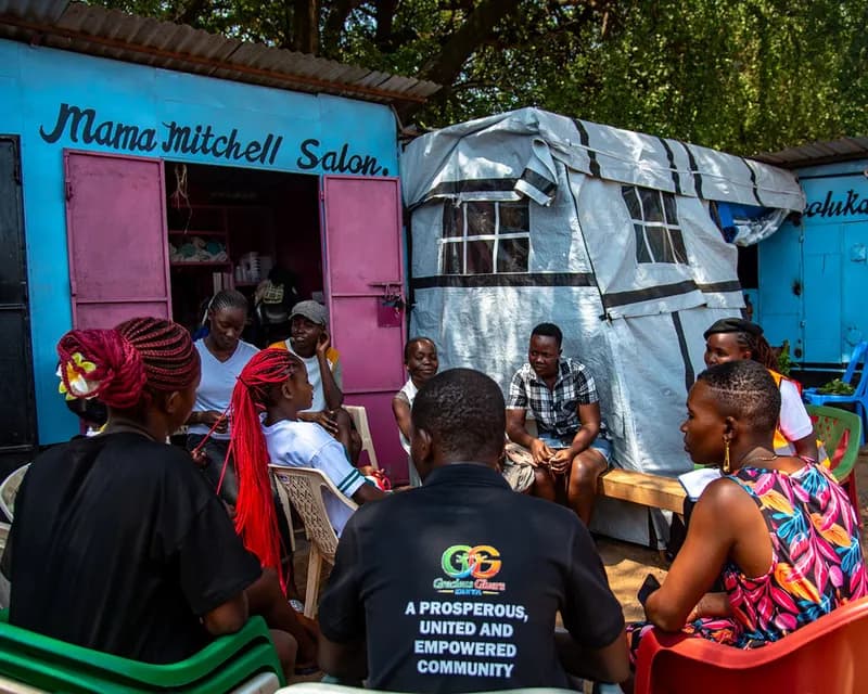Gracious Givers Kenya team members and young people from the community seated in a circle of chairs outside brightly painted market kiosks during a community dialogue session.