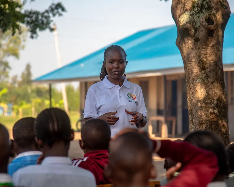 A Gracious Givers Kenya facilitator in a white polo speaks to a seated group of young people outside a blue-roofed school building in Nyando.