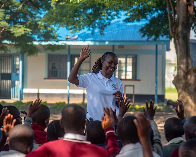 A smiling Gracious Givers Kenya facilitator raises her hand during an outdoor school session as the schoolchildren in front of her raise their hands to answer.