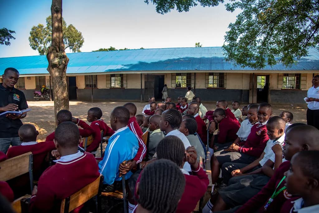 A Gracious Givers Kenya facilitator with a notebook speaks to a large group of students seated outdoors in front of their classroom block.