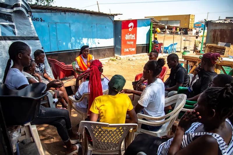 Community members and a Gracious Givers Kenya facilitator in an orange volunteer vest sit in a circle of plastic chairs for a dialogue session beside market kiosks in Nyando.