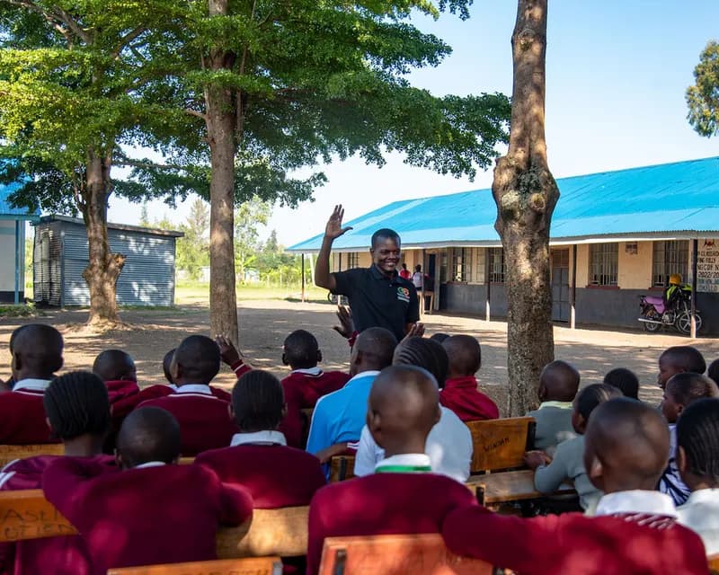 A Gracious Givers Kenya team member waves to rows of seated schoolchildren in a tree-shaded school compound with a blue-roofed classroom block behind.