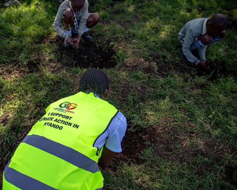 A volunteer in a yellow high-visibility vest reading “I support climate action in Nyando” kneels on the grass planting a tree seedling, with two young people planting alongside.